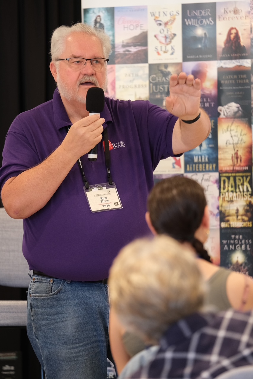 Speaker Rick Shaw, an older man with glasses and a purple polo, speaks into a handheld microphone while gesturing to an audience at a book event, with book covers in the background.