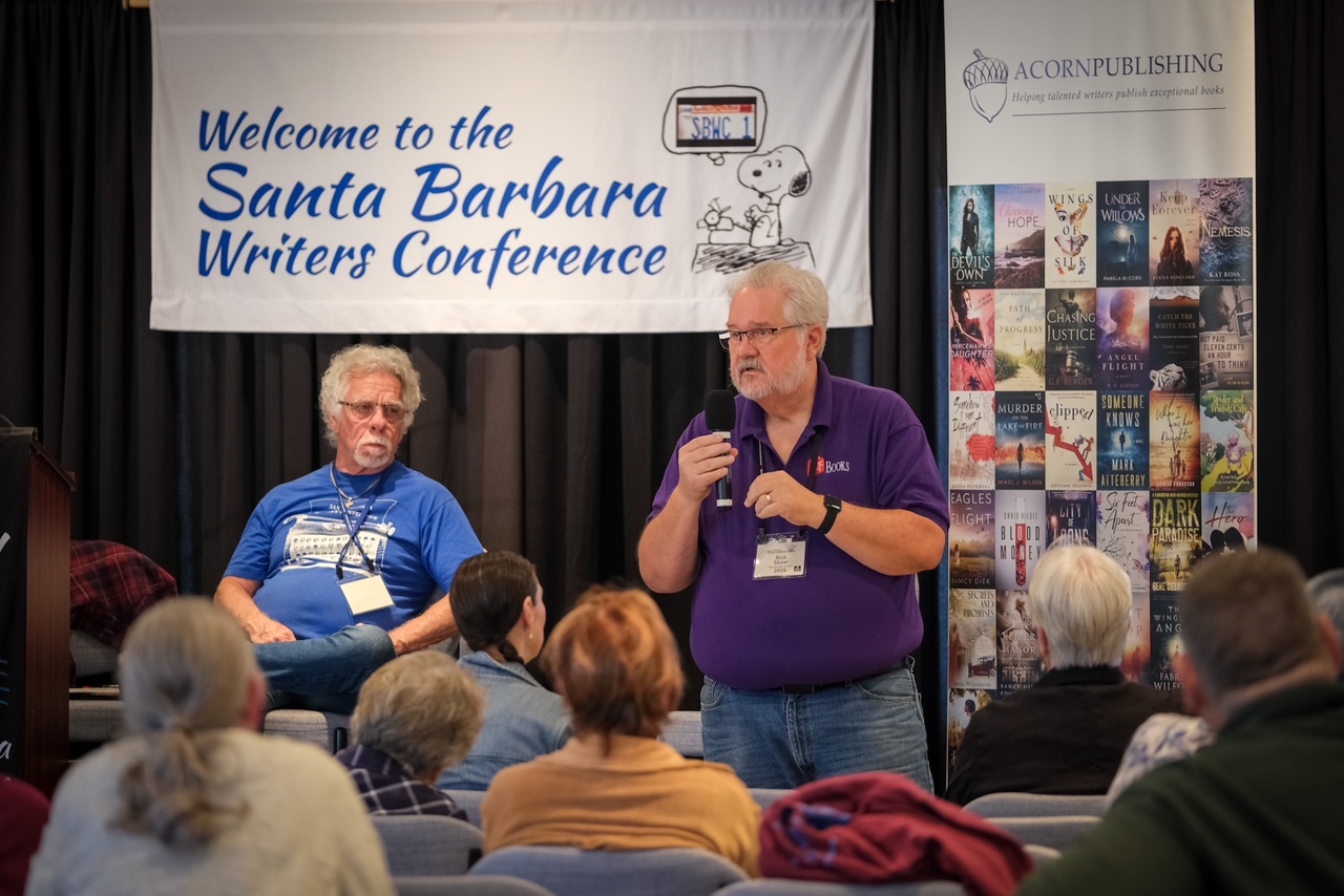 Speaker in a purple polo holds a microphone while addressing an audience at the Santa Barbara Writers Conference.